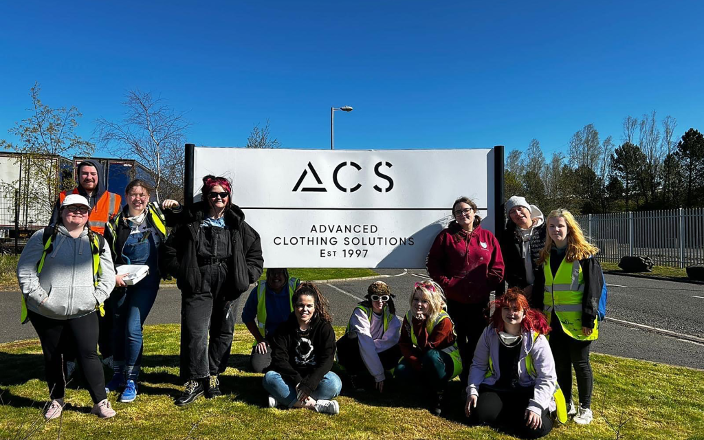 The group of New College Lanarkshire students sat around the ACS Sign in from of their Warehouse sitting in the sunlight with their mentor Mia McGregor-Webb