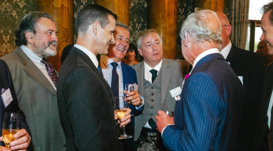 Michael Cusack, Head of Sustainability and business Transformation attends the reception celebrating the winners of the King's Award for Enterprise which is held by King Charles III, Michael is Centre frame mid discussion with King Charles, who has his back to the camera in a blue pinstripe suit. They are surrounded by fellow winners holding champagne flutes in upbeat conversation.